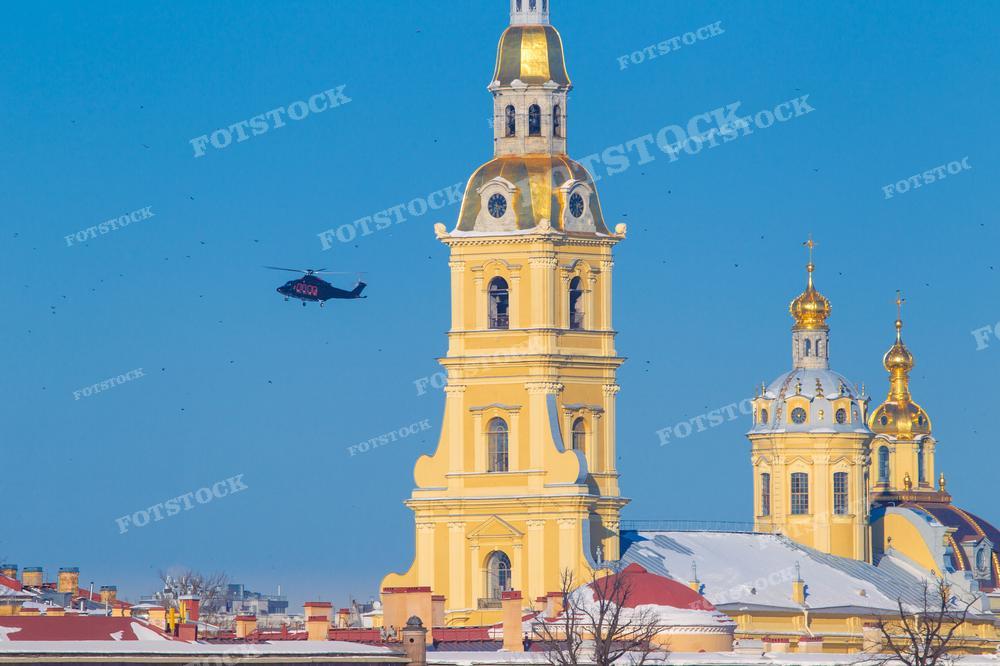На фотографии изображены золотые купола и колокольня православного собора Петропавловской крепости, на фоне голубого неба без облаков. В воздухе, на значительном расстоянии от здания, летит вертолет. Фотография может быть использована для иллюстрации туристических материалов о Санкт-Петербурге, статей о русской православной архитектуре, или как часть фотогалереи, демонстрирующей достопримечательности города. Также она подходит для новостных сообщений о событиях в городе, связанных с вертолетными полетами.
